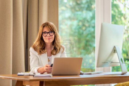 Photo for Cheerful smiling mid aged businesswoman sitting at table and using laptop for work. Confident professional female wearing white blazer and glasses. - Royalty Free Image