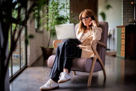 Photo for Full length of a mid aged woman wearing blazer and pants and sitting in an armchair. Confident mid aged woman using laptop for work or browsing on the internet. Home office. - Royalty Free Image
