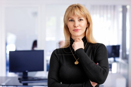 Photo for Close-up of blond haired woman standing at the office. Attractive businesswoman wearing turtleneck sweater while looking at camera and cheerful smiling. Copy space. - Royalty Free Image