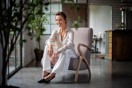 Photo for Full length of a beautiful woman wearing shirt and pants while sitting in an armchair at home. Cheerful smiling female relaxing by the window. - Royalty Free Image