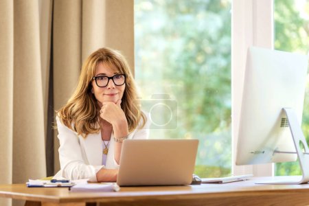 Téléchargez les photos : Joyeux sourire femme d'affaires d'âge moyen assis au bureau et utilisant un ordinateur portable pour le travail. Femme professionnelle confiante portant un blazer blanc et des lunettes. - en image libre de droit
