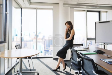 Téléchargez les photos : Toute la longueur d'une femme d'affaires attrayante debout au bureau et regardant pensif. Femme professionnelle exécutive ayant les cheveux longs brune et portant une robe noire. - en image libre de droit