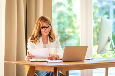 Foto de Mujer de negocios de mediana edad sentada en su escritorio y trabajando en su computadora portátil. Profesional femenino teniendo conferencia web. - Imagen libre de derechos
