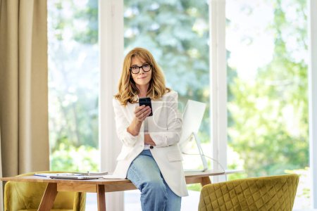 Photo for A middle aged woman sitting at a table and holding a phone in her hand. Attractive businesswoman looking at camera and smiling. She is wearing business casual and text messaging. - Royalty Free Image