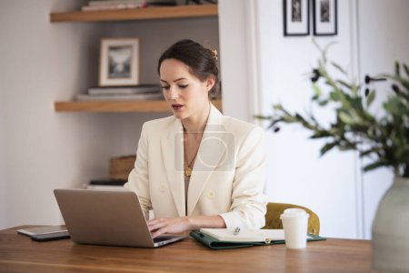 Photo for Attractive young business woman using laptop while sitting at desk and having video conference at the home office. Brunette haired female wearing white blazer. - Royalty Free Image