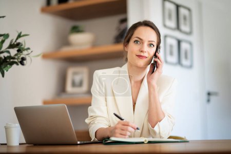 Foto de Mujer atractiva sentada en casa y trabajando. Mujer de negocios usando laptop y haciendo una llamada. Oficina en casa. - Imagen libre de derechos