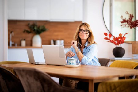Photo for Middle aged woman sitting at her dining room table and using earphone and laptop while having video call. Attractive female working at home. Home office. - Royalty Free Image
