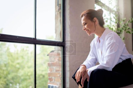 Foto de Atractiva mujer de mediana edad relajándose junto a la ventana y soñando despierto. Morena mujer de pelo sentado en una silla de salón y con camisa blanca y pantalones negros. - Imagen libre de derechos