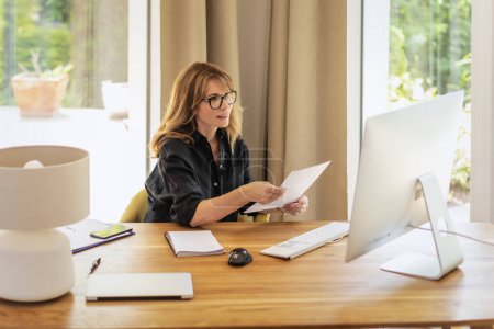 Téléchargez les photos : Femme d'affaires confiante utilisant l'ordinateur pour et ayant web conférence. Femme d'âge moyen assise à table et travaillant à domicile. - en image libre de droit
