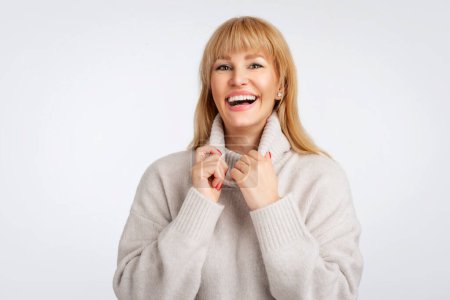 Foto de Mujer de mediana edad sonriente de pie sobre fondo blanco. Una mujer segura lleva suéter. Ella tiene el pelo rubio. - Imagen libre de derechos
