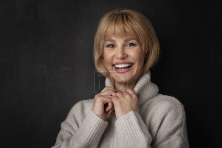 Foto de Sonriente mujer de mediana edad de pie sobre fondo negro. Una mujer segura lleva jersey de cuello alto. Ella tiene el pelo rubio. - Imagen libre de derechos