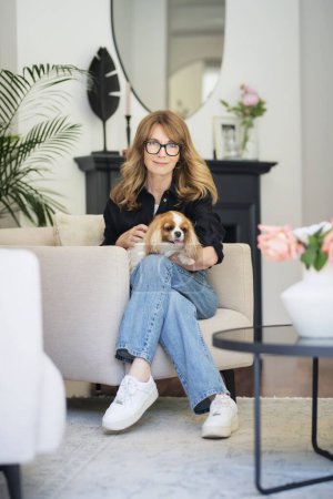 Photo for Full length of a middle-aged woman with blonde hair is sitting in an armchair at home with her cute dog. Attractive female wearing casual clothes. - Royalty Free Image