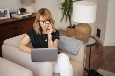 Foto de Retrato de mujer rubia sentada en un sillón en casa y usando laptop y smartphone. Atractiva mujer trabajando en casa. Oficina en casa. - Imagen libre de derechos
