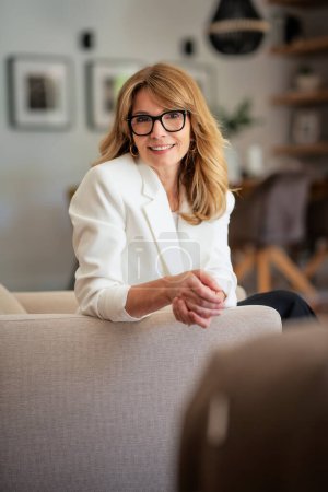 Photo for Mid aged attractive woman relaxing in an armchair at home. Blond haired female wearing eyeglasses and white blazer. - Royalty Free Image