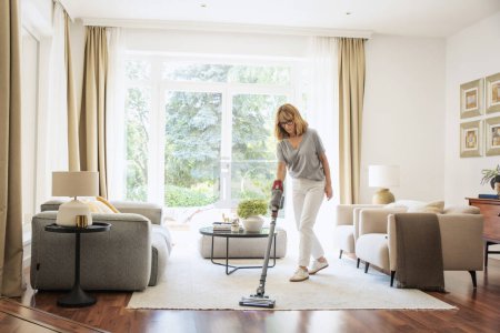 Téléchargez les photos : Femme d'âge moyen nettoyage tapis avec aspirateur à la maison. Femme fait des travaux ménagers dans le salon. - en image libre de droit
