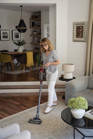 Photo for Mid aged woman cleaning carpet with vacuum cleaner at home. Female is doing housework in living room. - Royalty Free Image