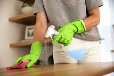 Photo for Close-up of a woman's hand wiping the dining table at home. Confident female wearing rubber gloves and using cleaning liquid. - Royalty Free Image