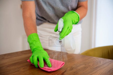 Foto de Primer plano de la mano de una mujer limpiando la mesa del comedor en casa. Mujer segura que usa guantes de goma y líquido de limpieza. - Imagen libre de derechos