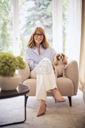 Photo for Full length of an attractive blond haired woman relaxing in armchair at home with her cute puppy. Mid aged female looking at camera and cheerful smiling. Attractive woman wearing blue shirt and white pants. - Royalty Free Image
