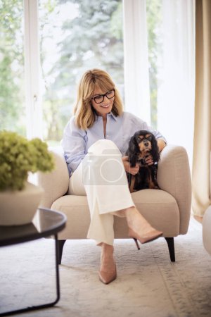 Foto de Longitud completa de una atractiva mujer rubia que se relaja en sillón en casa con su lindo cachorro. Mujer de mediana edad mirando a su perro y sonriendo alegre. Mujer atractiva con camisa azul y pantalones blancos. - Imagen libre de derechos