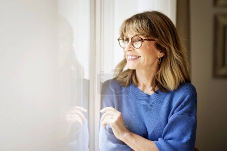 Photo for Portrait of a beautiful middle-aged woman standing at the window. Blond haired female wearing blue shirt and eyewear and looking out. Copy space. - Royalty Free Image