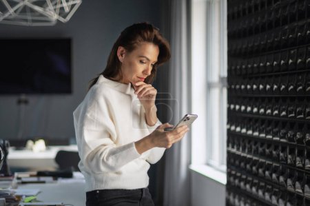 Foto de Una mujer de negocios de mediana edad de pie en la oficina y utilizando un teléfono inteligente. Atractiva hembra mirando reflexivo y mensajes de texto. - Imagen libre de derechos