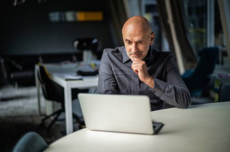 Photo for Shot of serious thoughtful businessman sitting at table with laptop at the office in late evening and working. - Royalty Free Image