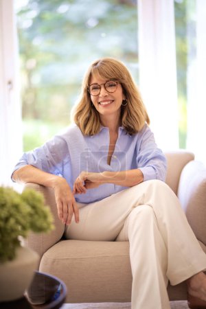 Foto de Retrato de una hermosa mujer con el pelo rubio está sentado en un sillón en casa. Mujer riéndose vistiendo camisa azul y pantalones blancos. - Imagen libre de derechos