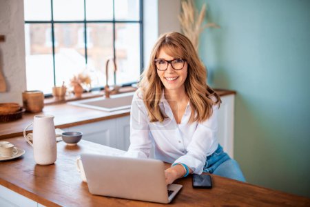 Photo for A middle-aged woman standing at the kitchen counter in the morning and using a laptop. Confident female wearing casual clothes and having video call. Home office. - Royalty Free Image