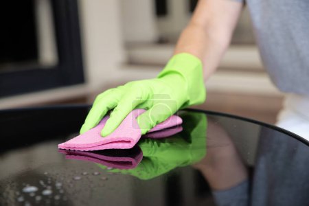 Foto de Primer plano de la mano de una mujer limpiando la mesa de café en casa. Mujer segura que usa guantes de goma y líquido de limpieza mientras limpia en casa. - Imagen libre de derechos