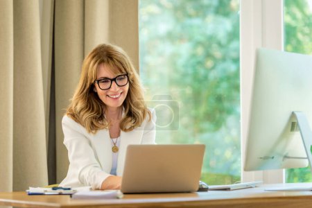 Foto de Mujer de negocios rubia de pelo sentado en su escritorio y trabajando en su computadora portátil. Mujer profesional que usa gafas y blazer blanco. Oficina en casa. - Imagen libre de derechos