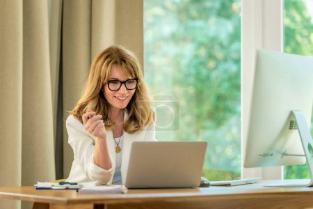Photo for Blonde haired businesswoman sitting at her desk and working on her laptop. Professional female wearing eyewear and white blazer. Home office. - Royalty Free Image