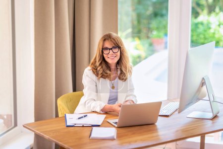 Photo for Blonde haired businesswoman sitting at her desk and working on her laptop. Professional female wearing eyewear and white blazer. - Royalty Free Image