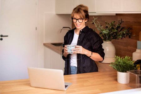 Photo for Blond haired mid aged woman standing in the kitchen at home and using notebook. Smiling female having video call and drinking tea. Home office. - Royalty Free Image