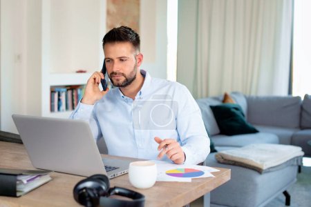 Foto de Pensando hombre de negocios trabajando desde casa mientras está sentado detrás de su portátil y teniendo una llamada. Oficina en casa. - Imagen libre de derechos