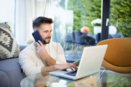 Photo for Close-up of handsome man sitting at home and using a laptop and mobile phone. Mid aged male wearing casual clothes and working at home. Home office. - Royalty Free Image