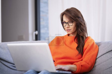 Photo for Serious faced brunette haired woman sitting on sofa at home and using laptop for work. Attractive female wearing orange sweater. Home office. - Royalty Free Image