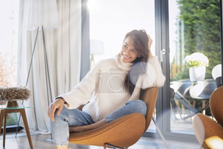 Photo for Cheerful smiling brunette haired woman relaxing in an armchair at home. Attractive female wearing casual clothes and looking away. - Royalty Free Image