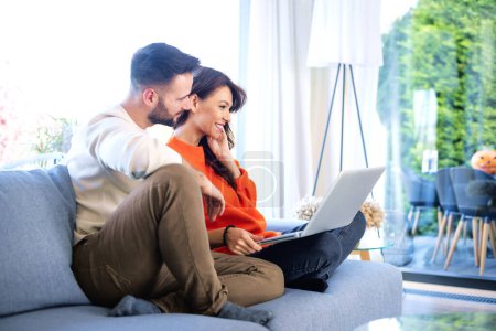 Photo for Shot of a young couple using a laptop on their sofa at home. Happy woman and man wearing casual clothes and browsing on the internet or having video call. - Royalty Free Image