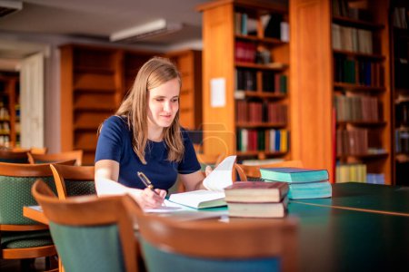 Téléchargez les photos : Une jeune femme malvoyante assise et étudiant à la bibliothèque universitaire. Jeune femme utilisant des livres et des notes. - en image libre de droit