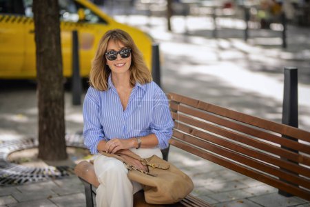 Photo for Attractive woman with blonde hair is sitting on a bench in the street. Mid aged female looking at camera and smiling. - Royalty Free Image