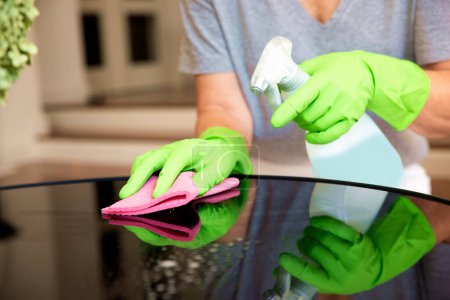 Foto de Primer plano de la mano de una mujer limpiando la mesa de café en casa. Mujer segura que usa guantes de goma y líquido de limpieza mientras limpia en casa. - Imagen libre de derechos
