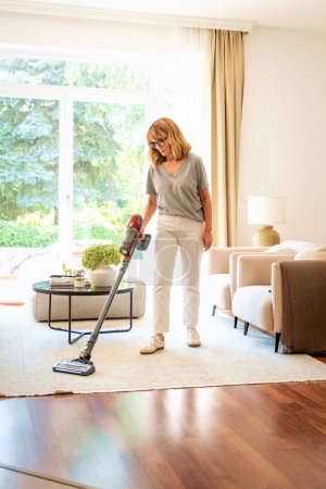Photo for Mid aged woman cleaning carpet with vacuum cleaner at home. Female doing housework in living room. Full length shot. - Royalty Free Image