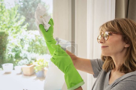 Photo for Smiling mid aged woman cleaning and wiping window with spray bottle at home. Confident female wearing rubber gloves and casual clothes. - Royalty Free Image