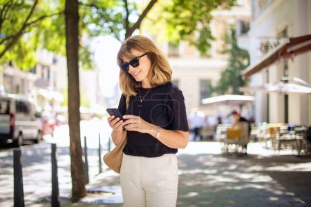 Téléchargez les photos : Femme d'âge moyen debout dans la rue de la ville et utilisant un smartphone. Attrayant féminin portant des vêtements décontractés et la messagerie texte. - en image libre de droit
