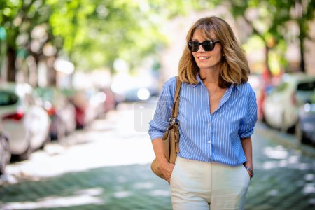 Foto de Hermosa mujer de mediana edad caminando por la calle de la ciudad. Mujer rubia vistiendo camisa azul con pantalones blancos. - Imagen libre de derechos