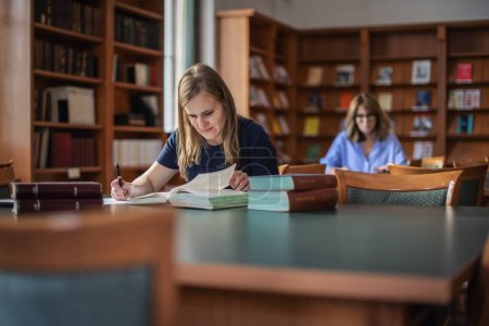 Photo for A visually impaired young woman sitting and studying in the university library. Young female using books and making notes. - Royalty Free Image