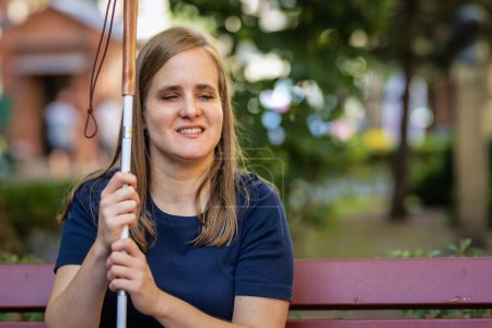 Foto de Primer plano de una mujer con discapacidad visual sosteniendo un bastón blanco y sentada en un banco de la ciudad. Mujer sonriente vistiendo ropa casual. - Imagen libre de derechos