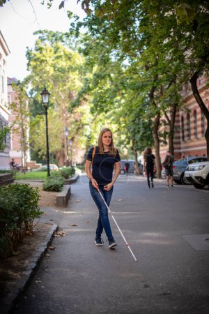 Foto de Retrato de mujer ciega con bastón blanco caminando por la calle. Una mujer con discapacidad visual que viste ropa casual y usa su bastón para cruzar la calle. Disparo completo. - Imagen libre de derechos