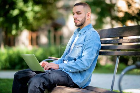 Téléchargez les photos : Jeune homme utilisant un ordinateur portable pour étudier ou travailler à l'extérieur. Jeune homme portant des vêtements décontractés et assis sur le banc dans le parc public. - en image libre de droit